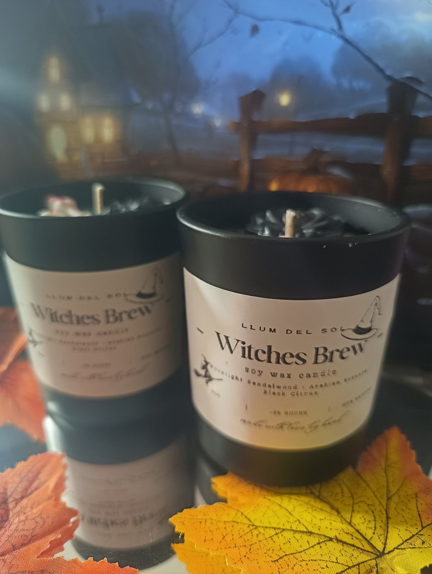 Two 'Witches Brew' candles with a blurred background of trees and lights.