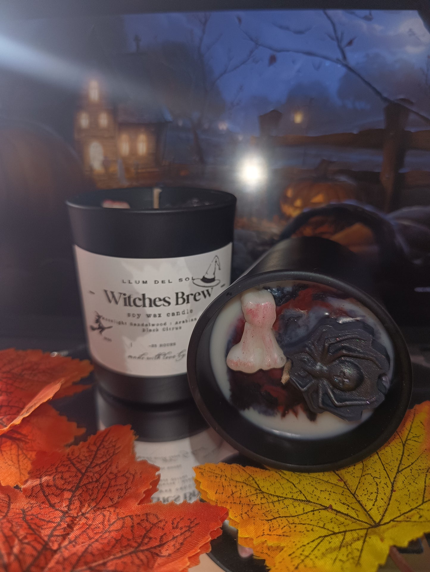 Witches Brew candle with decorative elements on a reflective surface with autumn leaves.