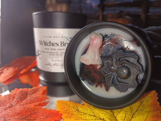 Candle with spider design and 'Witches Brew' label, surrounded by autumn leaves.